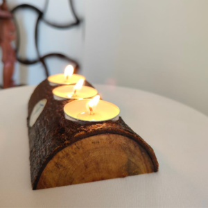 A handcrafted three-hole natural wood log tea light holder with three lit candles and a small blue circular emblem on the dark bark surface.